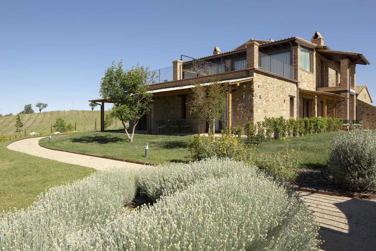 Elegant stone house surrounded by lush greenery and lavender plants, featuring a curved pathway leading to the entrance under a clear blue sky. | Only on SopranoVillas