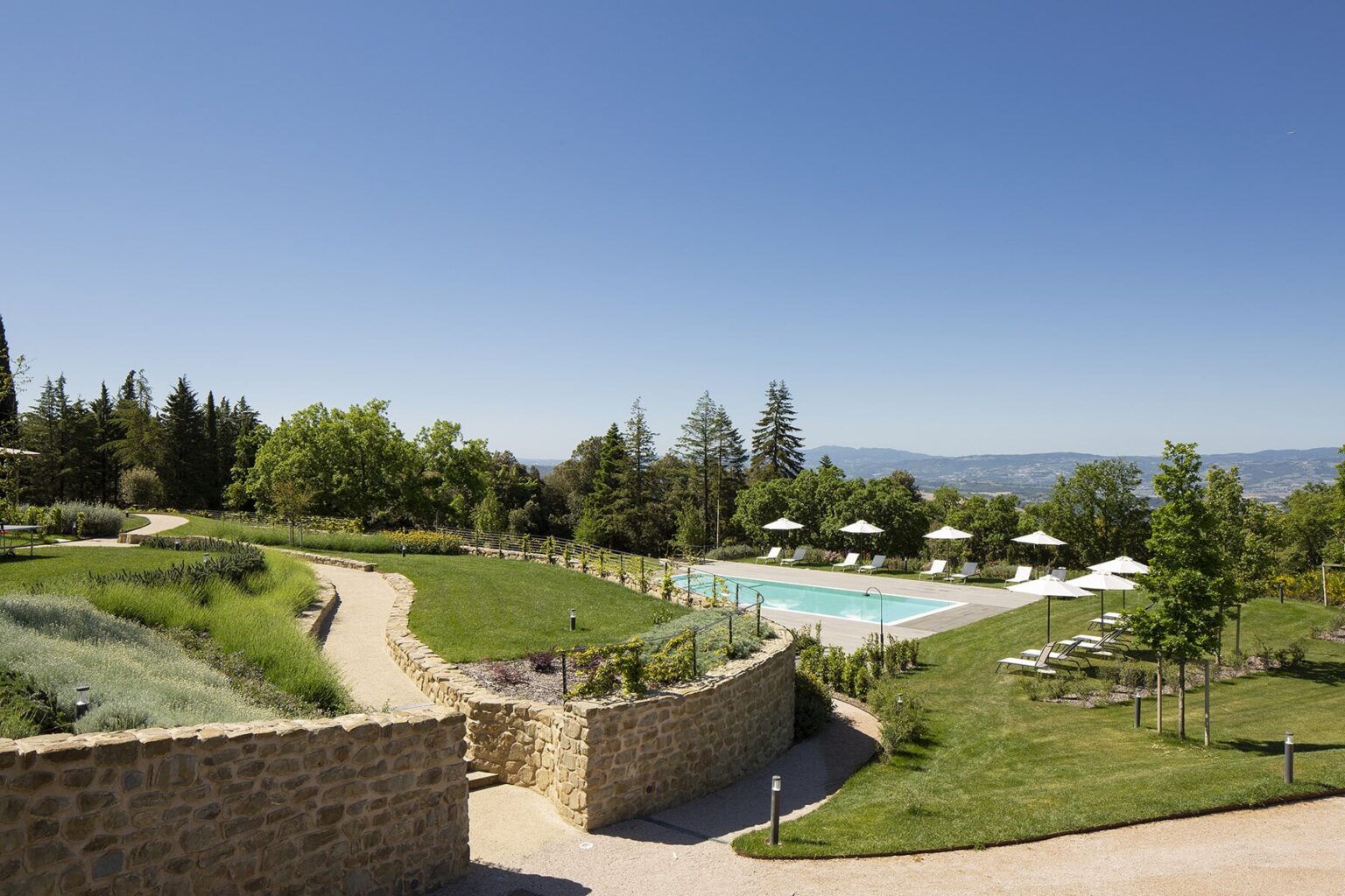 Scenic view of a luxury resort featuring a swimming pool surrounded by lush greenery, sun loungers, and trees under a clear blue sky. Ideal for relaxation and outdoor activities. | Only on SopranoVillas