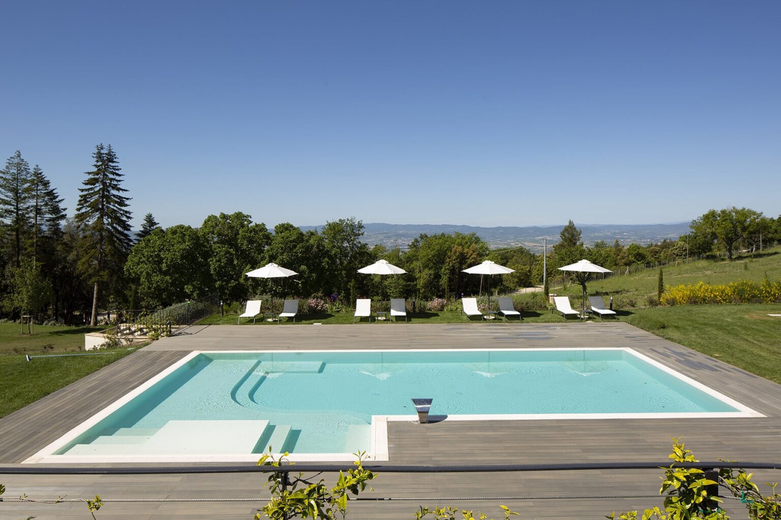 Modern outdoor swimming pool surrounded by lounge chairs and umbrellas, set in a lush green landscape under a clear blue sky. Ideal for relaxation and leisure activities. | Only on SopranoVillas
