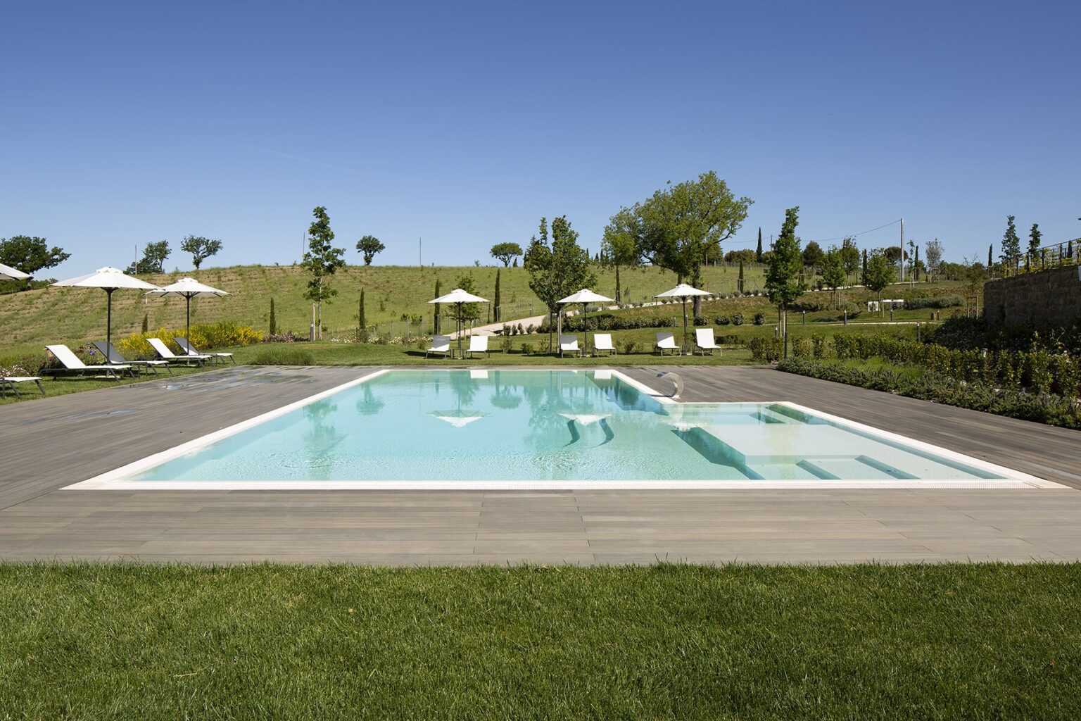 Modern outdoor swimming pool surrounded by lush green grass and sun loungers, with umbrellas providing shade, set against a clear blue sky and landscaped hills in the background. | Only on SopranoVillas
