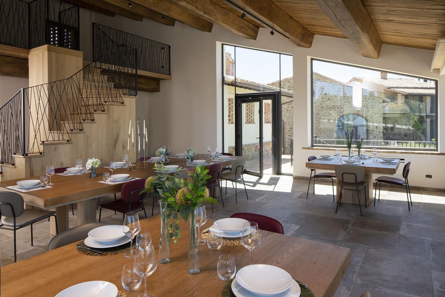 Modern dining area with wooden tables set for a meal, featuring elegant tableware and floral centerpieces, large windows offering natural light, and a stylish staircase in the background. | Only on SopranoVillas