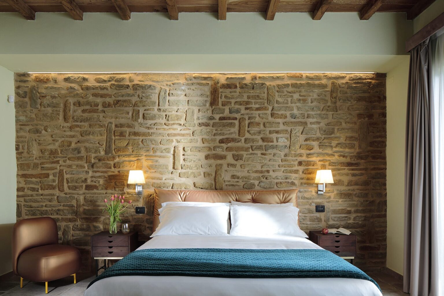 Cozy bedroom featuring a stone accent wall, a comfortable bed with white linens and a teal throw blanket, modern bedside lamps, and a stylish armchair, enhancing the inviting atmosphere of the space. | Only on SopranoVillas