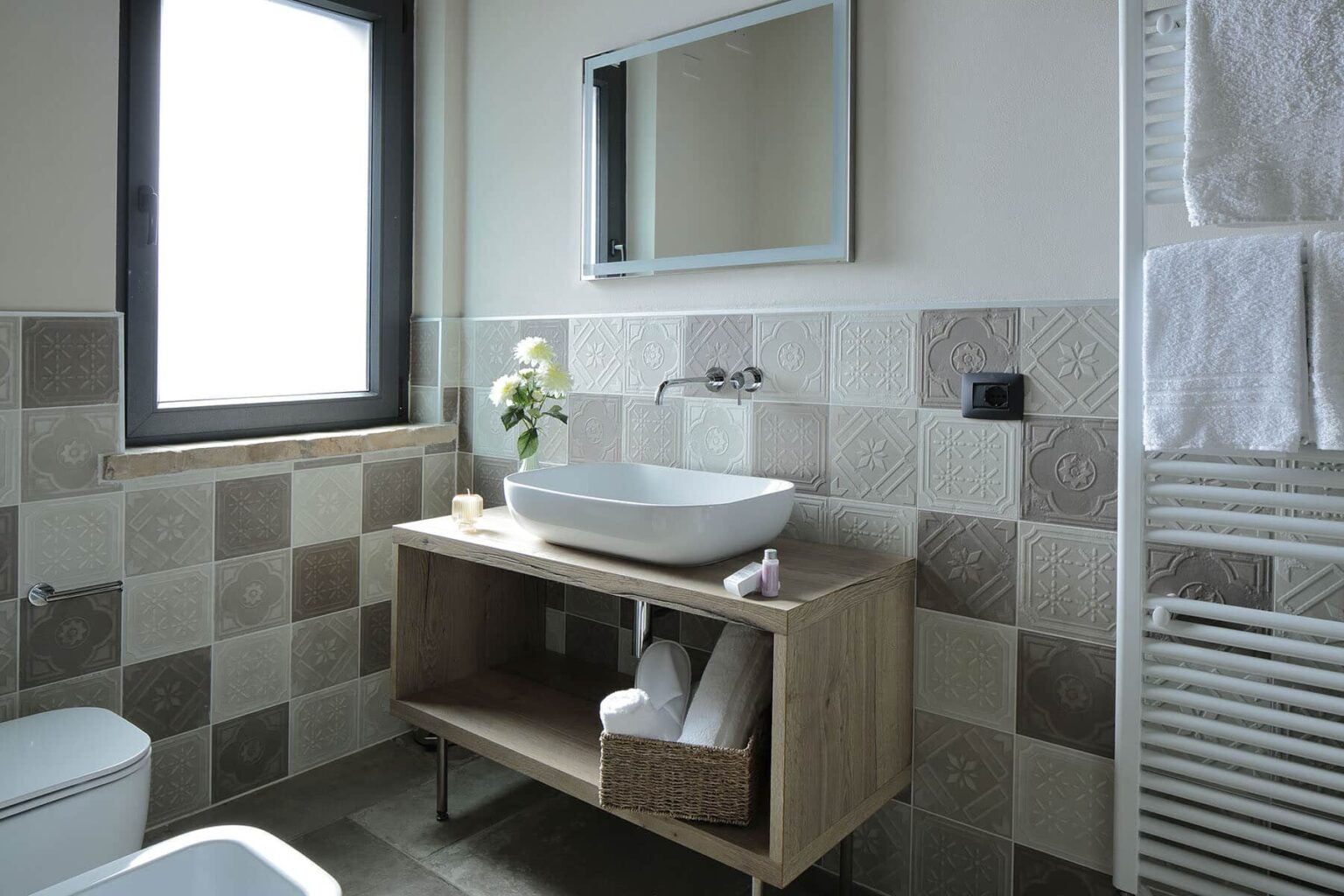 Modern bathroom interior featuring a stylish wooden vanity with a sink, neatly arranged towels, a wall-mounted mirror, and a window for natural light. The walls are adorned with a mix of textured tiles, enhancing the contemporary design. | Only on SopranoVillas