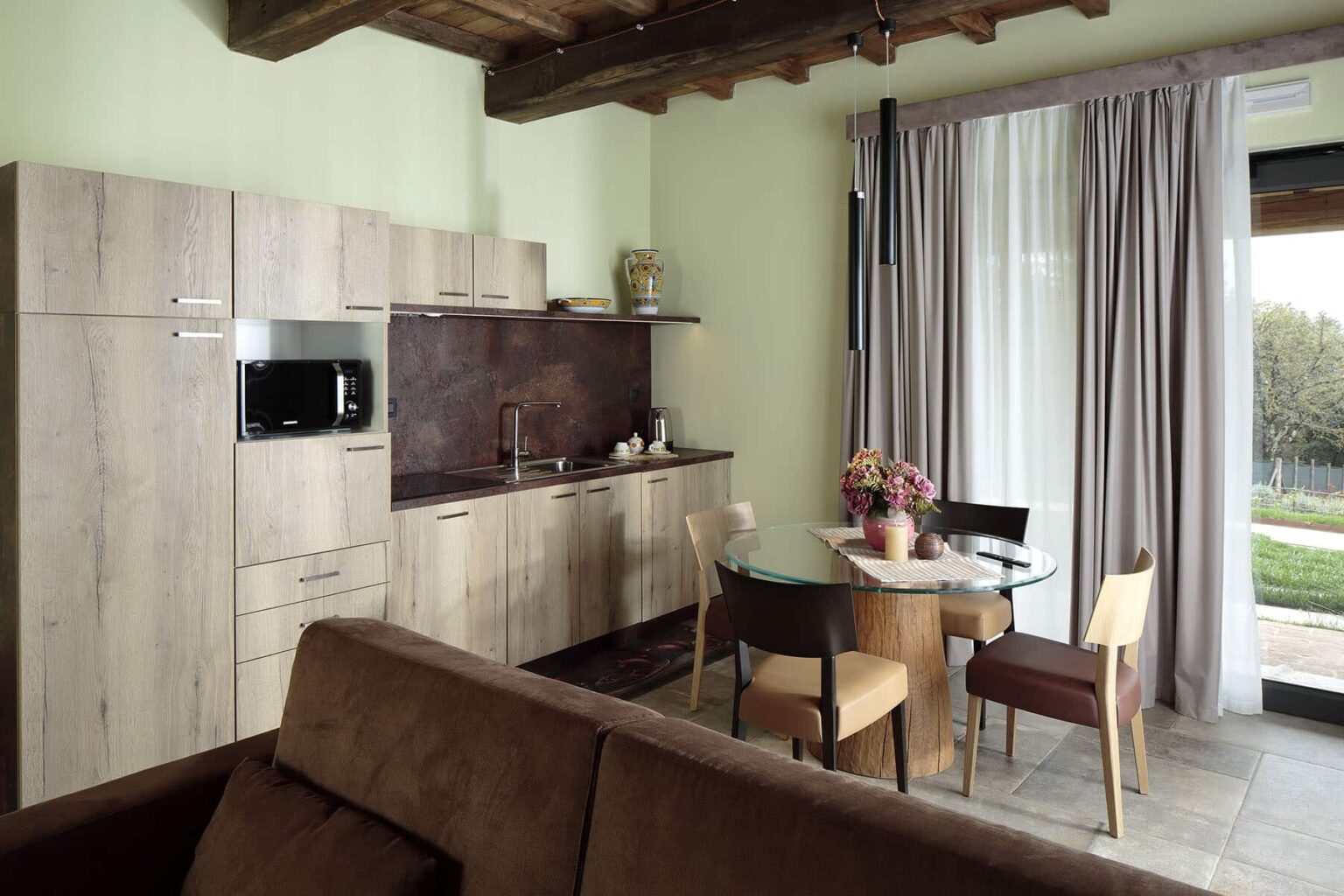 Modern kitchen and dining area featuring wooden cabinets, a round dining table with chairs, and large windows letting in natural light. The decor includes neutral tones and contemporary design elements, creating a cozy and inviting atmosphere. | Only on SopranoVillas