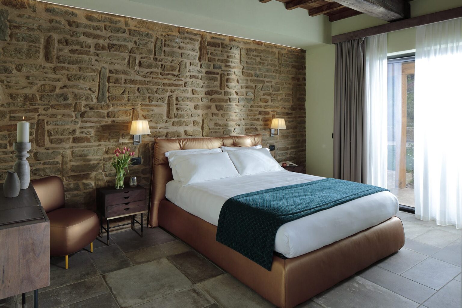 Modern hotel room featuring a comfortable queen-sized bed with a teal throw blanket, set against a textured stone wall. The room includes stylish bedside lamps, a wooden nightstand, and large windows allowing natural light to brighten the space. | Only on SopranoVillas