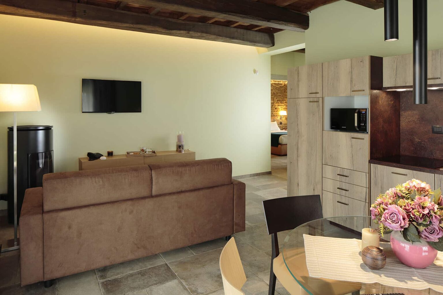 Modern living room and kitchen area featuring a brown sofa, wooden furniture, and a vase of flowers, with soft lighting and a flat-screen TV on the wall. | Only on SopranoVillas