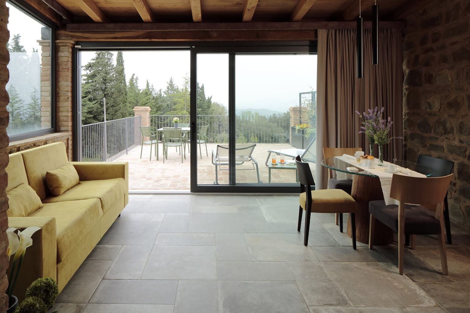 Modern living room interior featuring a yellow sofa, dining table with chairs, and large glass doors opening to a scenic outdoor view. Natural light floods the space through the windows, highlighting the rustic stone walls and contemporary design elements. | Only on SopranoVillas