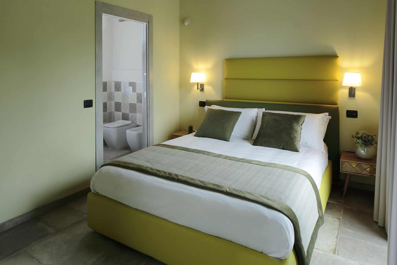 Cozy bedroom featuring a queen-sized bed with green and white bedding, modern wall sconces, and a view of an en-suite bathroom. The room has a light green color scheme and stylish decor, ideal for relaxation. | Only on SopranoVillas