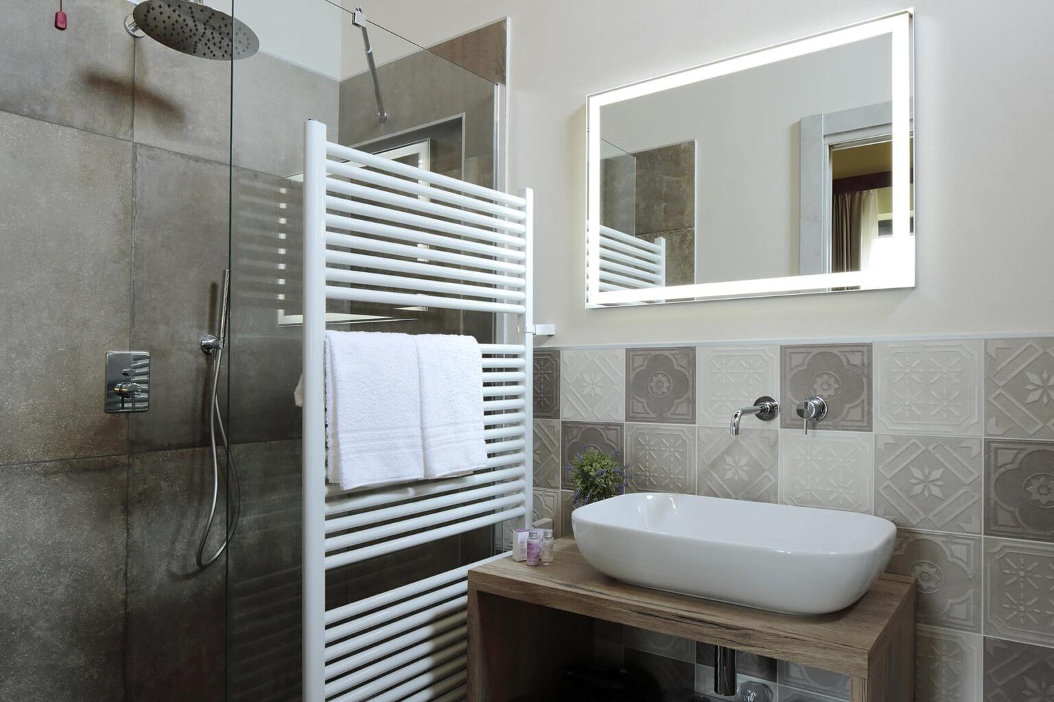 Modern bathroom design featuring a glass shower, a white heated towel rack, and a stylish sink on a wooden countertop. The walls are adorned with textured tiles, and a sleek mirror enhances the contemporary aesthetic. | Only on SopranoVillas
