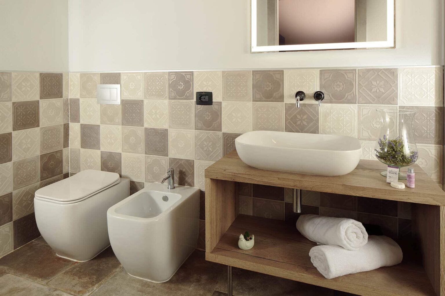 Modern bathroom design featuring a bidet and toilet, a stylish countertop basin, and a wooden shelf with neatly rolled towels. The walls are adorned with a mosaic of beige and brown tiles, complemented by a rectangular mirror. | Only on SopranoVillas