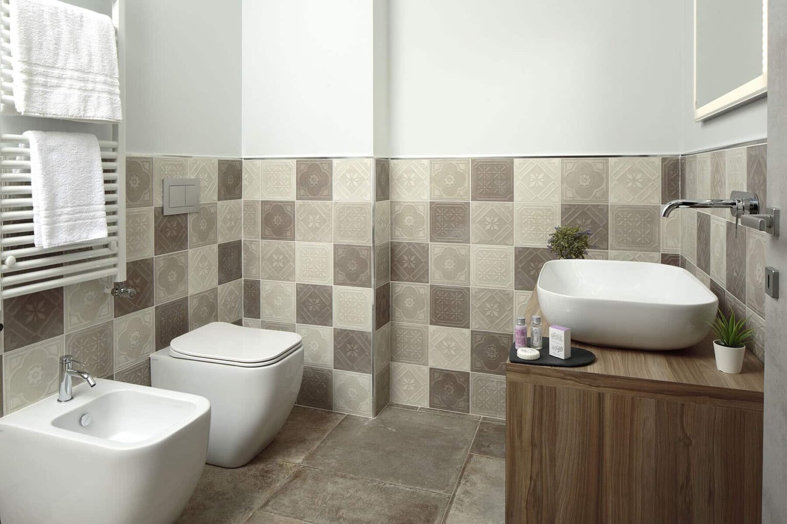 Modern bathroom featuring a stylish design with patterned tile walls, a wall-mounted sink, a bidet, and a wooden vanity. The space is well-lit and includes a towel rack, creating a clean and inviting atmosphere. | Only on SopranoVillas