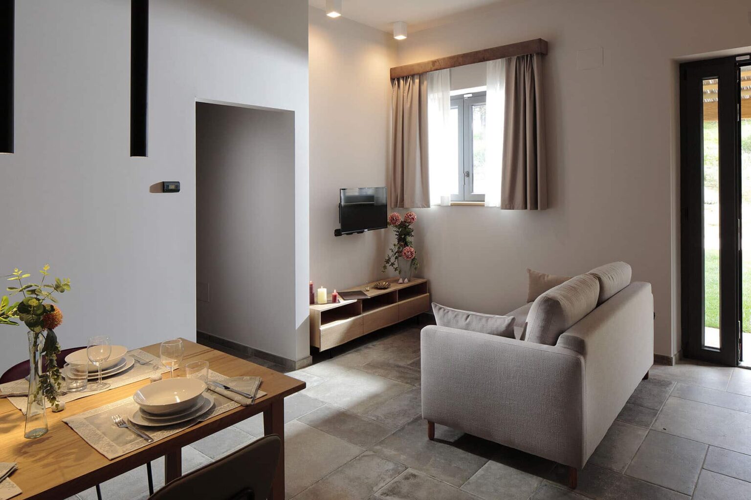 Modern living room with a cozy sofa, wooden TV stand, and dining table set for two. Natural light filters through the window with curtains, illuminating the stylish decor and stone flooring. | Only on SopranoVillas