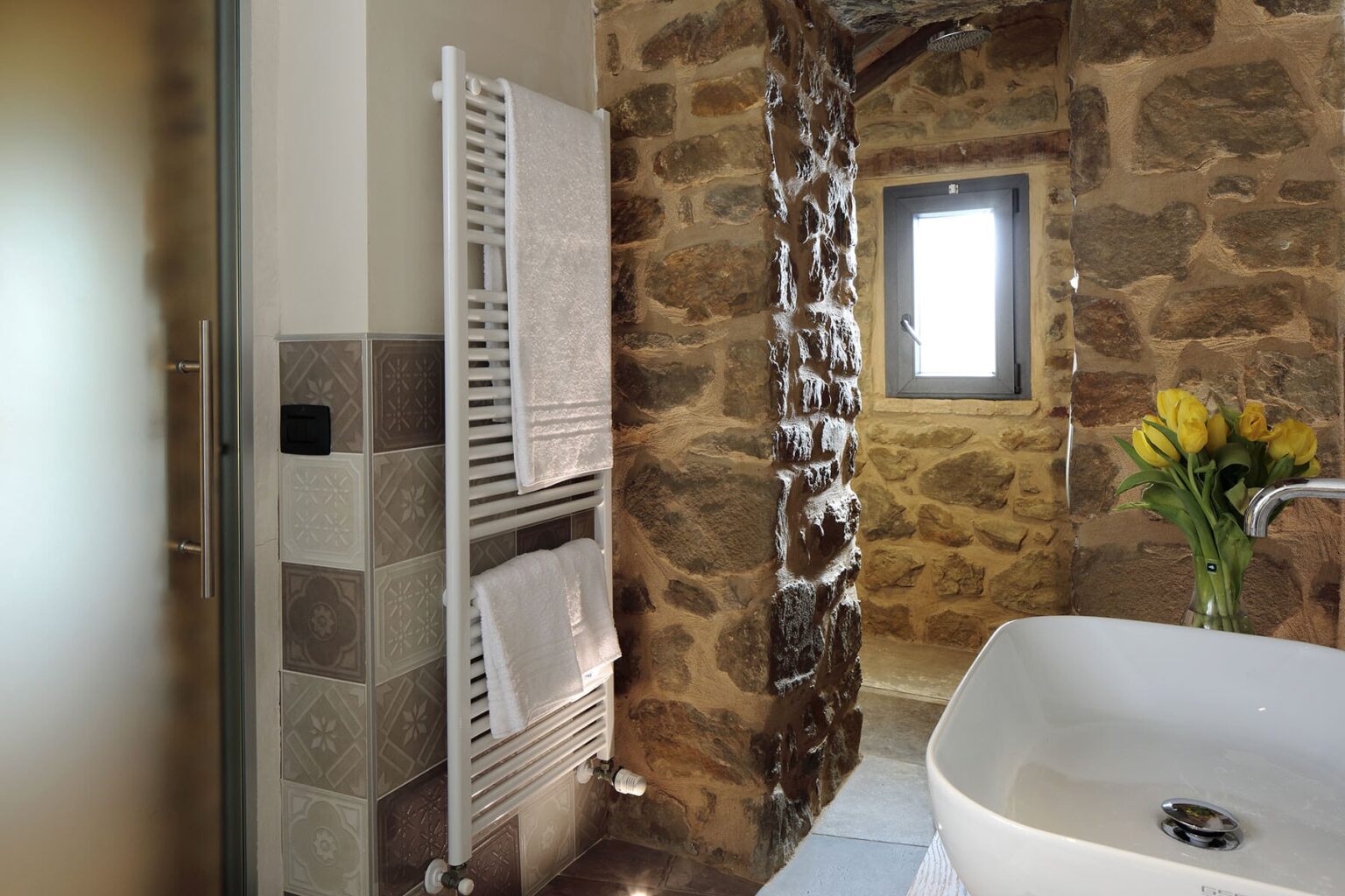 Modern bathroom featuring a stone wall, stylish bathtub, and a towel radiator, with a window allowing natural light and a vase of yellow flowers for a touch of color. | Only on SopranoVillas