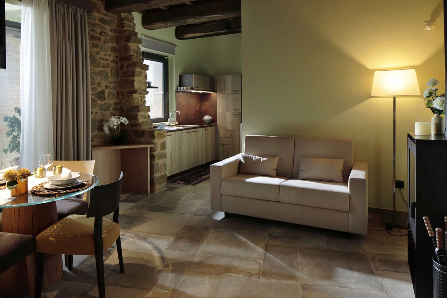 Cozy living room with a beige sofa, wooden dining table set with breakfast, and a modern kitchen in the background. Soft lighting and stone walls create a warm atmosphere. | Only on SopranoVillas