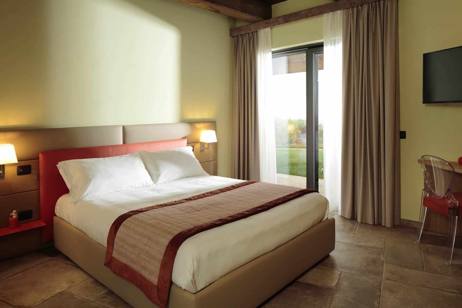 Modern hotel room featuring a queen-sized bed with stylish bedding, large windows allowing natural light, and a flat-screen TV, ideal for relaxation and comfort during your stay. | Only on SopranoVillas