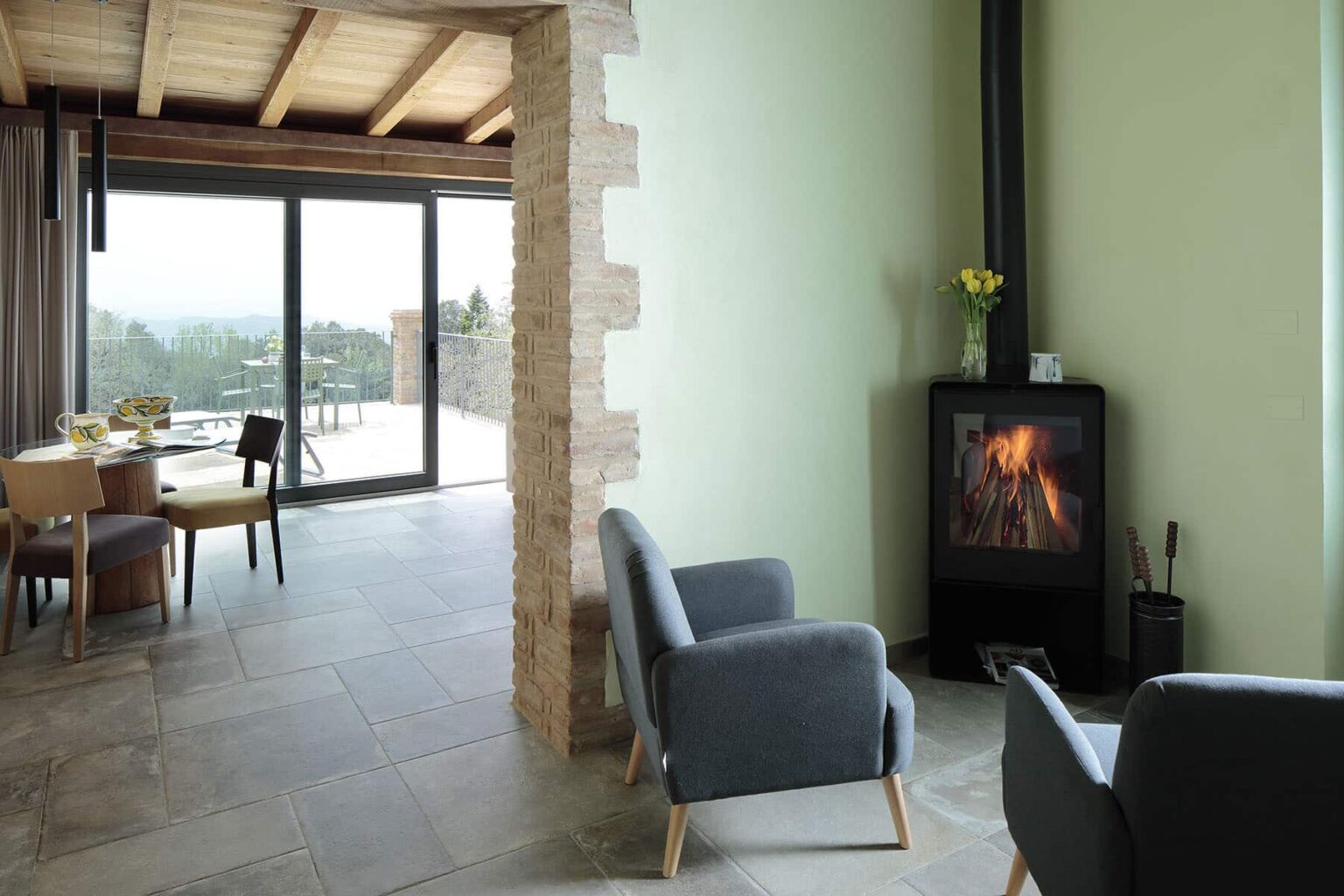 Modern living room featuring a cozy wood stove, stylish gray armchairs, and large glass doors opening to a scenic view. The interior showcases a blend of natural stone and soft green walls, creating a warm and inviting atmosphere. | Only on SopranoVillas