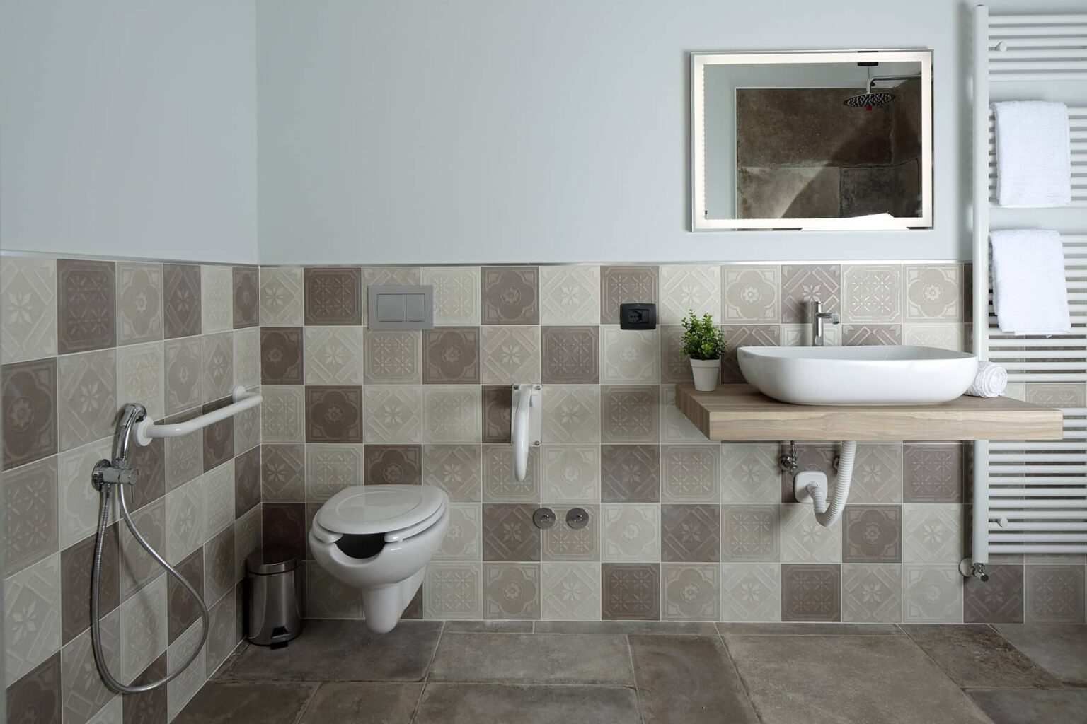 Modern bathroom design featuring a wall-mounted sink, a contemporary toilet, and stylish tiled walls in neutral tones. The space includes a mirror, hand towels, and a small plant for added decor. | Only on SopranoVillas