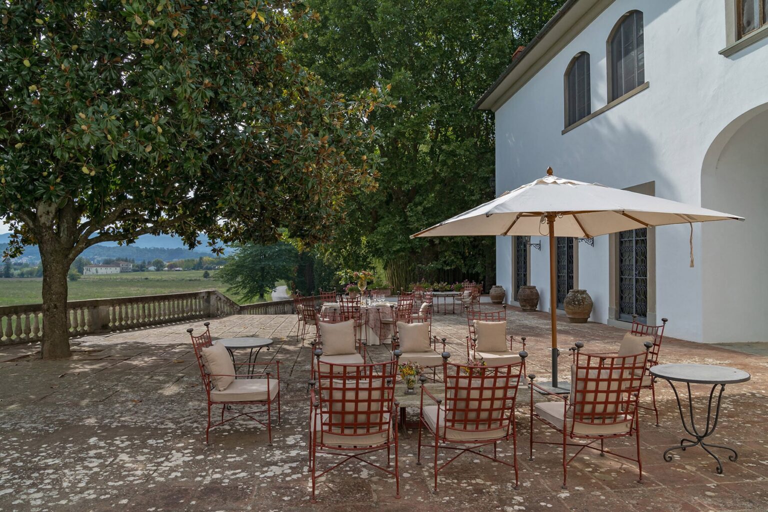 Outdoor dining area with a large umbrella and wooden chairs set under a tree, adjacent to a white building, offering a scenic view of the countryside. Perfect for gatherings and events. | Only on SopranoVillas