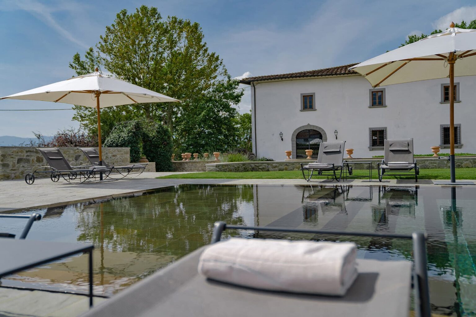 Luxurious outdoor pool area featuring lounge chairs and umbrellas, with a beautiful white villa in the background surrounded by greenery. Perfect for relaxation and leisure in a serene setting. | Only on SopranoVillas