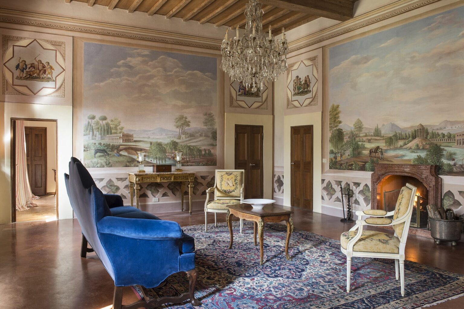 Elegant living room with vintage decor featuring a blue velvet sofa, ornate chandelier, and hand-painted murals depicting serene landscapes. The room is enhanced by a classic rug and wooden furnishings, creating a sophisticated atmosphere. | Only on SopranoVillas