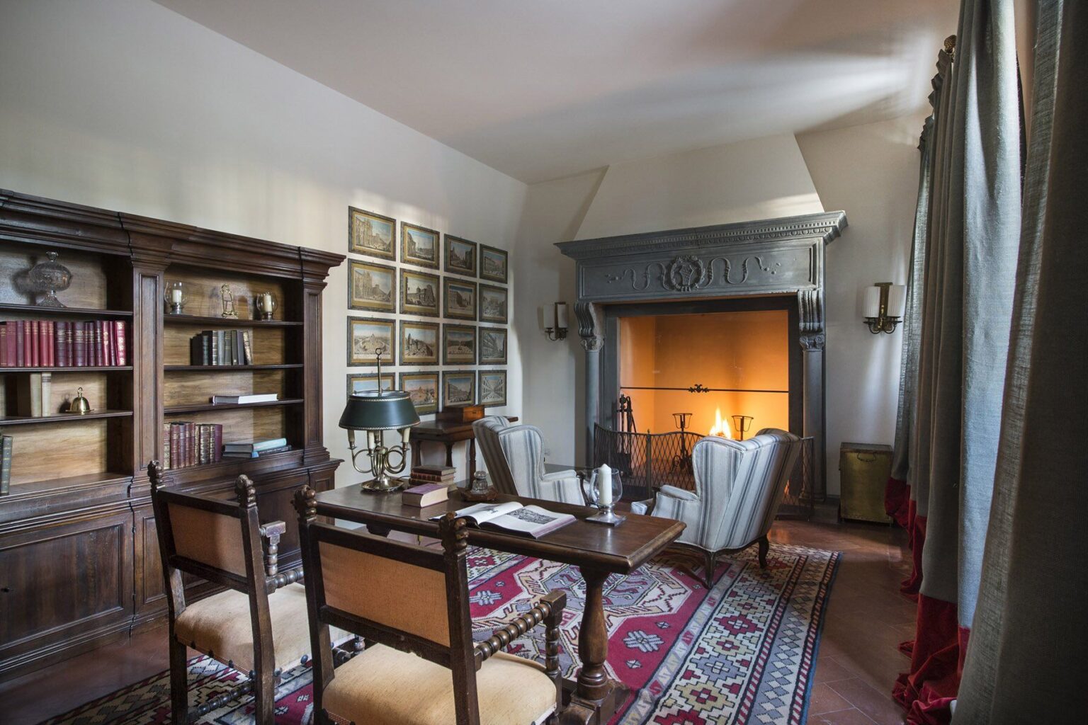 Cozy home office interior featuring a wooden desk, two chairs, a bookshelf filled with books, and a fireplace. The room is decorated with warm lighting and elegant curtains, creating an inviting atmosphere. | Only on SopranoVillas