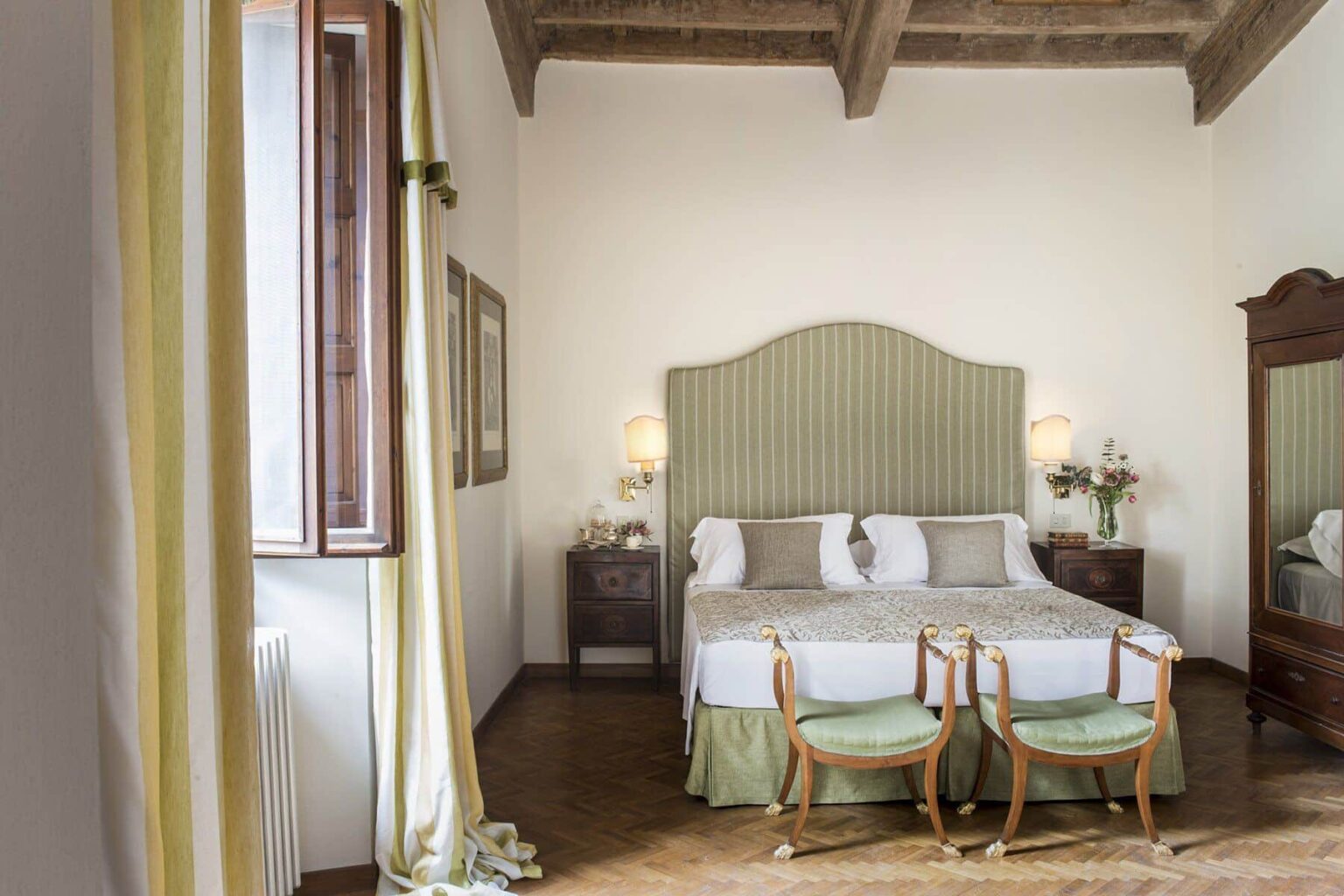 Cozy hotel room featuring a large bed with a green striped headboard, two wooden chairs, elegant decor, and natural light from a window with yellow curtains. | Only on SopranoVillas