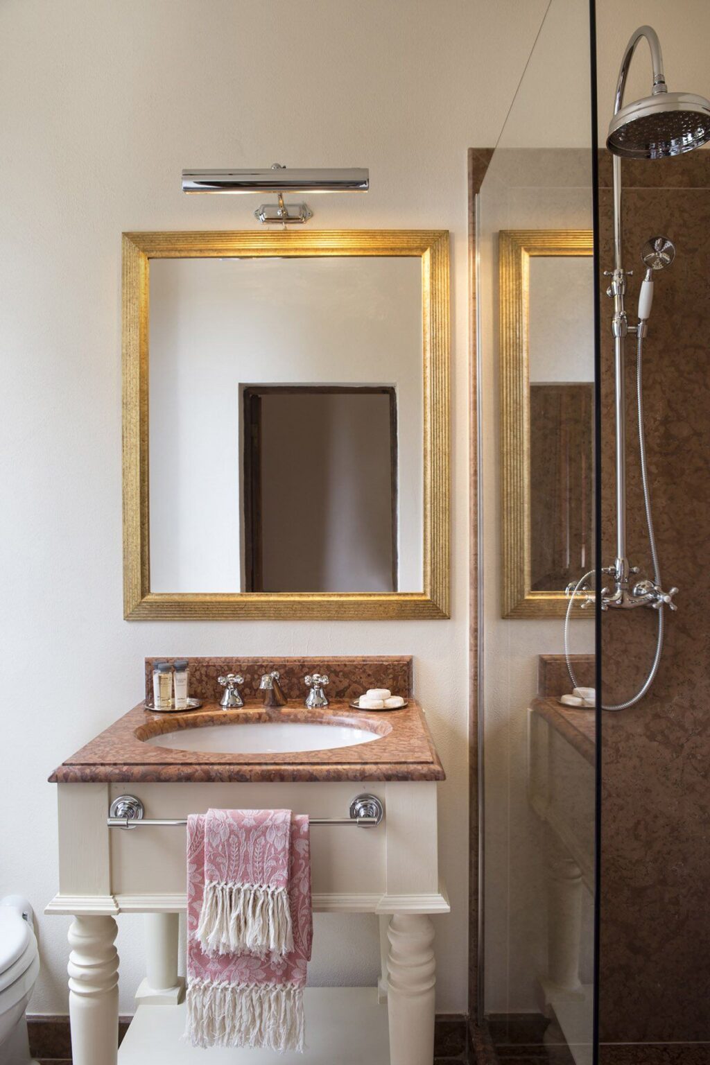 Elegant bathroom interior featuring a large framed mirror, a stylish sink with gold-toned faucets, and a glass shower. The warm color palette includes marble countertops and neutral walls, creating a serene atmosphere. | Only on SopranoVillas