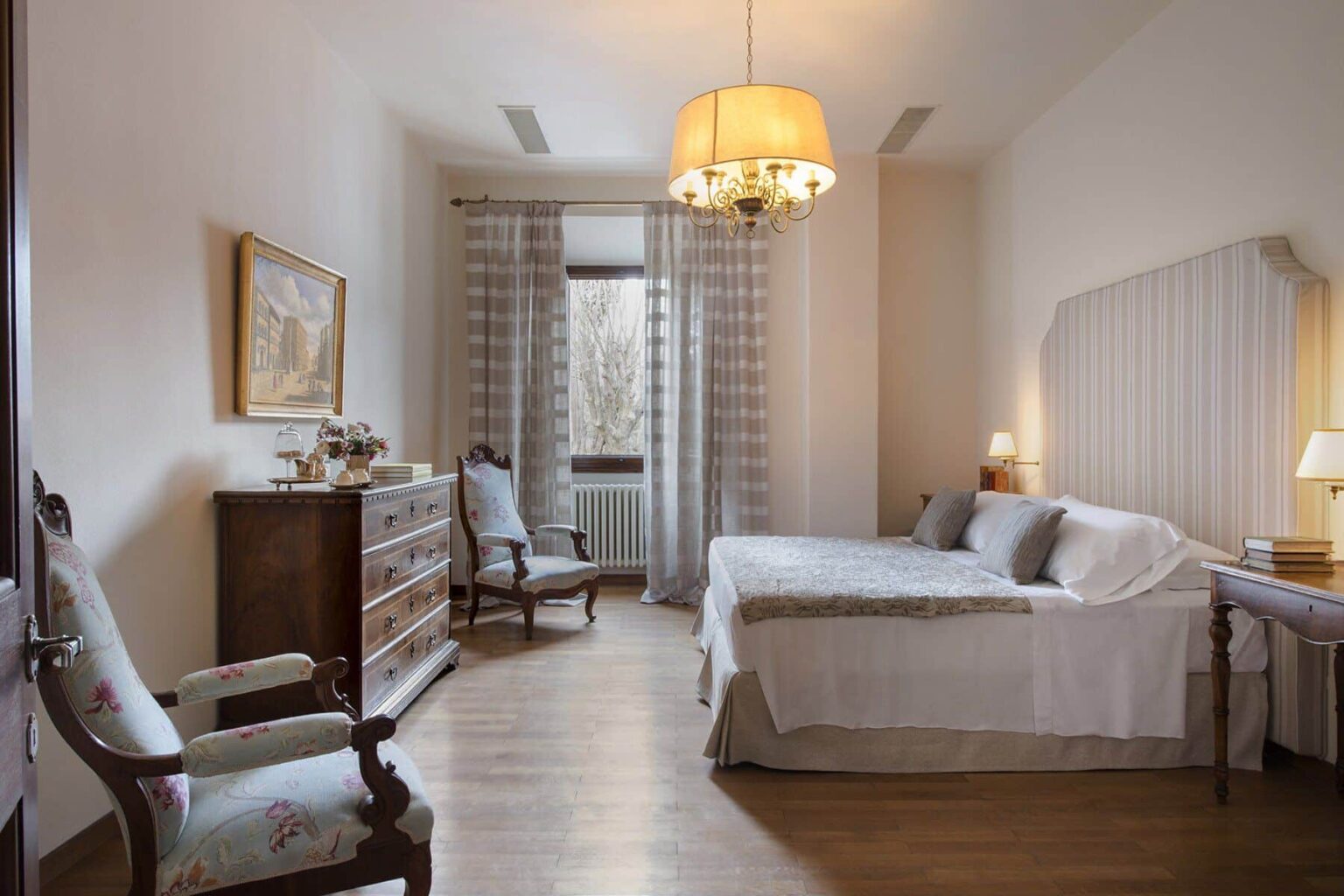 Cozy and elegant bedroom featuring a comfortable double bed, a wooden dresser, and stylish armchairs. Natural light streams through the large window adorned with sheer curtains, complemented by a decorative chandelier. Ideal for relaxation and restful sleep. | Only on SopranoVillas