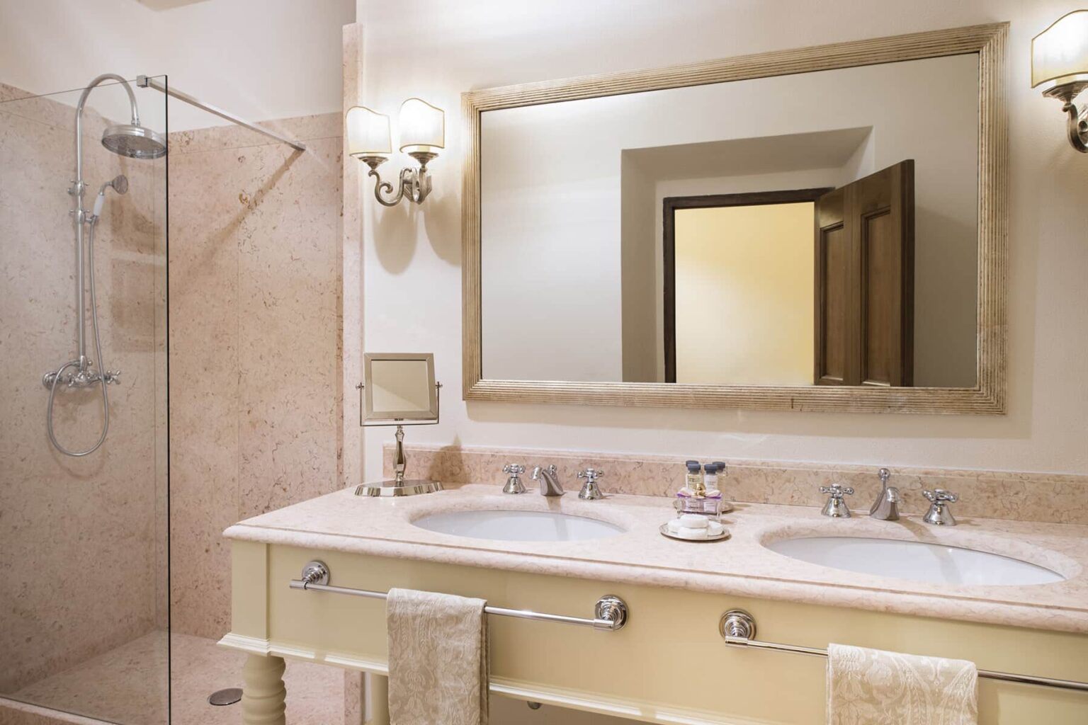 Elegant bathroom interior featuring a double sink vanity, large mirror, and a glass shower. The design includes soft lighting fixtures and a neutral color palette, creating a sophisticated atmosphere. | Only on SopranoVillas