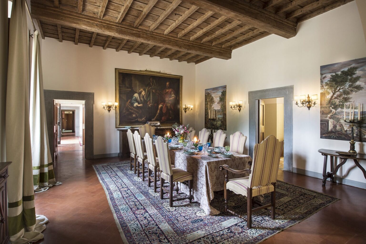 Elegant dining room featuring a long wooden table set for a meal, surrounded by plush chairs. The room boasts a traditional wooden ceiling, ornate wall sconces, and large paintings, creating a sophisticated atmosphere. A richly patterned rug adds warmth to the space. | Only on SopranoVillas