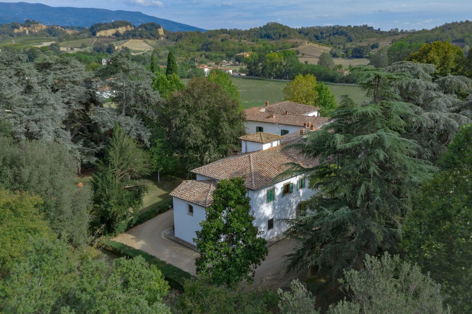 Aerial view of a picturesque villa surrounded by lush greenery and rolling hills, showcasing a blend of modern and traditional architecture in a serene landscape. | Only on SopranoVillas