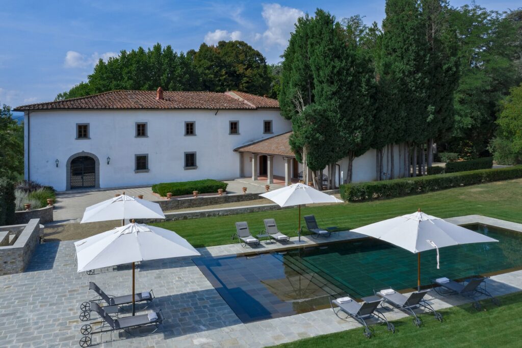 Aerial view of a luxurious villa with a swimming pool, surrounded by lush greenery and sun loungers under white umbrellas. The elegant architecture features a combination of modern and traditional design elements, set in a tranquil outdoor setting. | Only on SopranoVillas