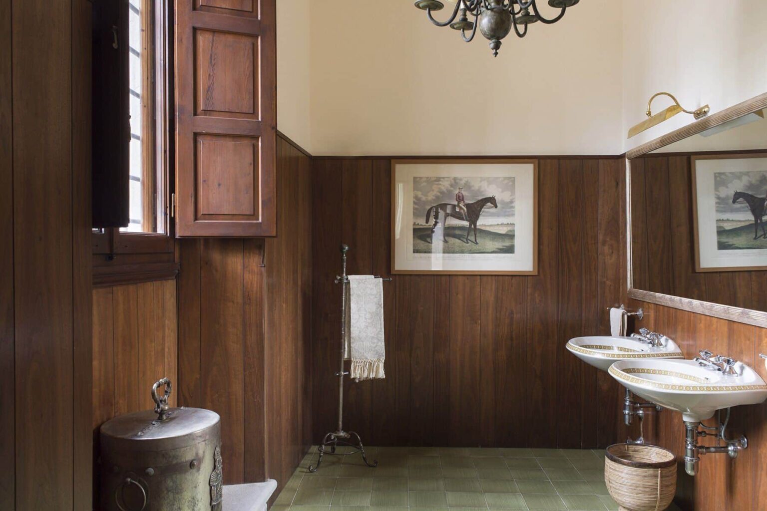 Vintage bathroom interior featuring wooden paneling, two sinks, a mirror, and a framed horse artwork on the wall. The space is illuminated by a chandelier and includes a towel rack and a trash can. | Only on SopranoVillas