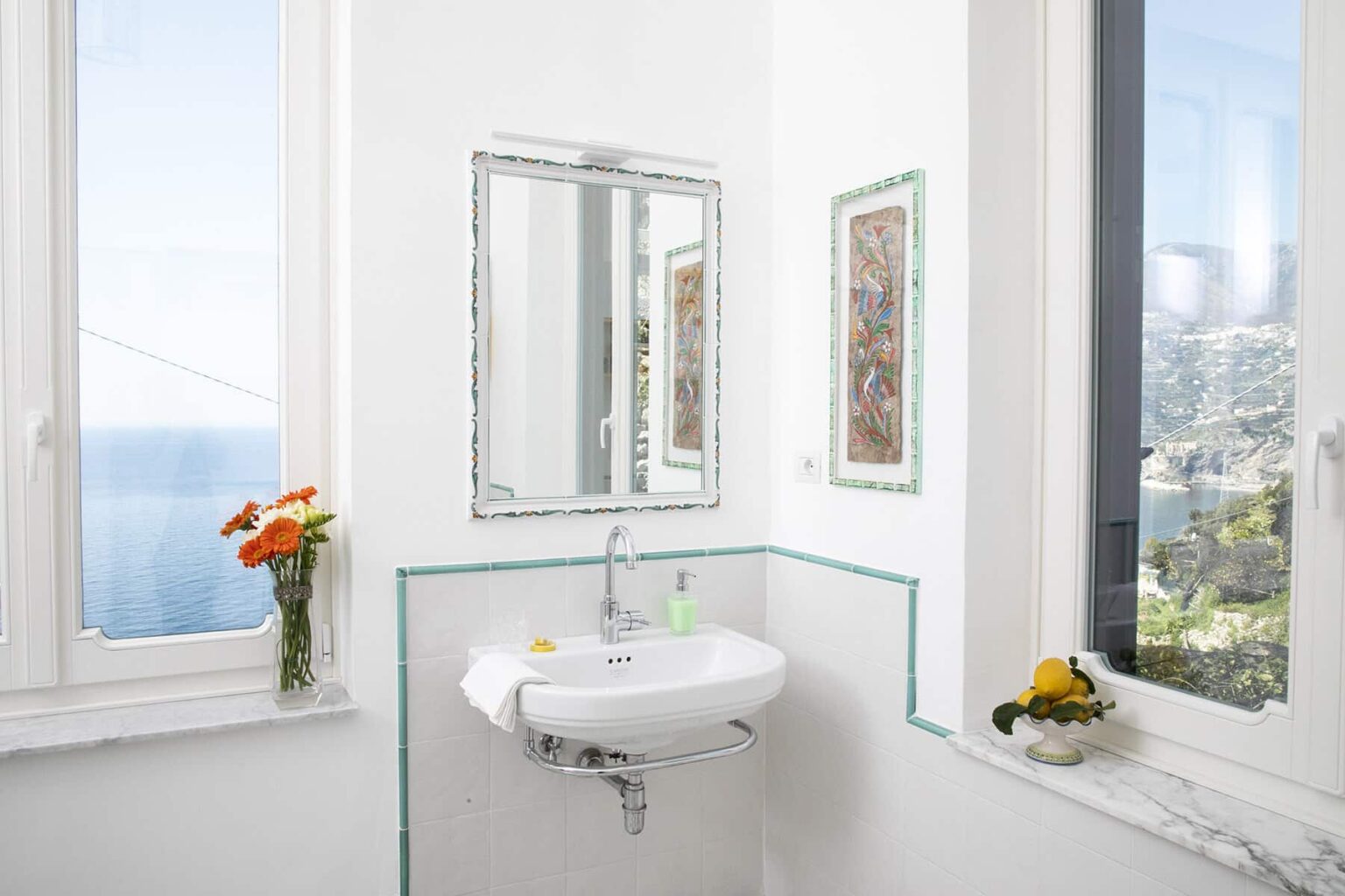 Bright and airy bathroom featuring a modern sink, large mirror, and scenic ocean view through expansive windows. Fresh flowers and decorative elements add a touch of elegance to the serene atmosphere. | Only on SopranoVillas
