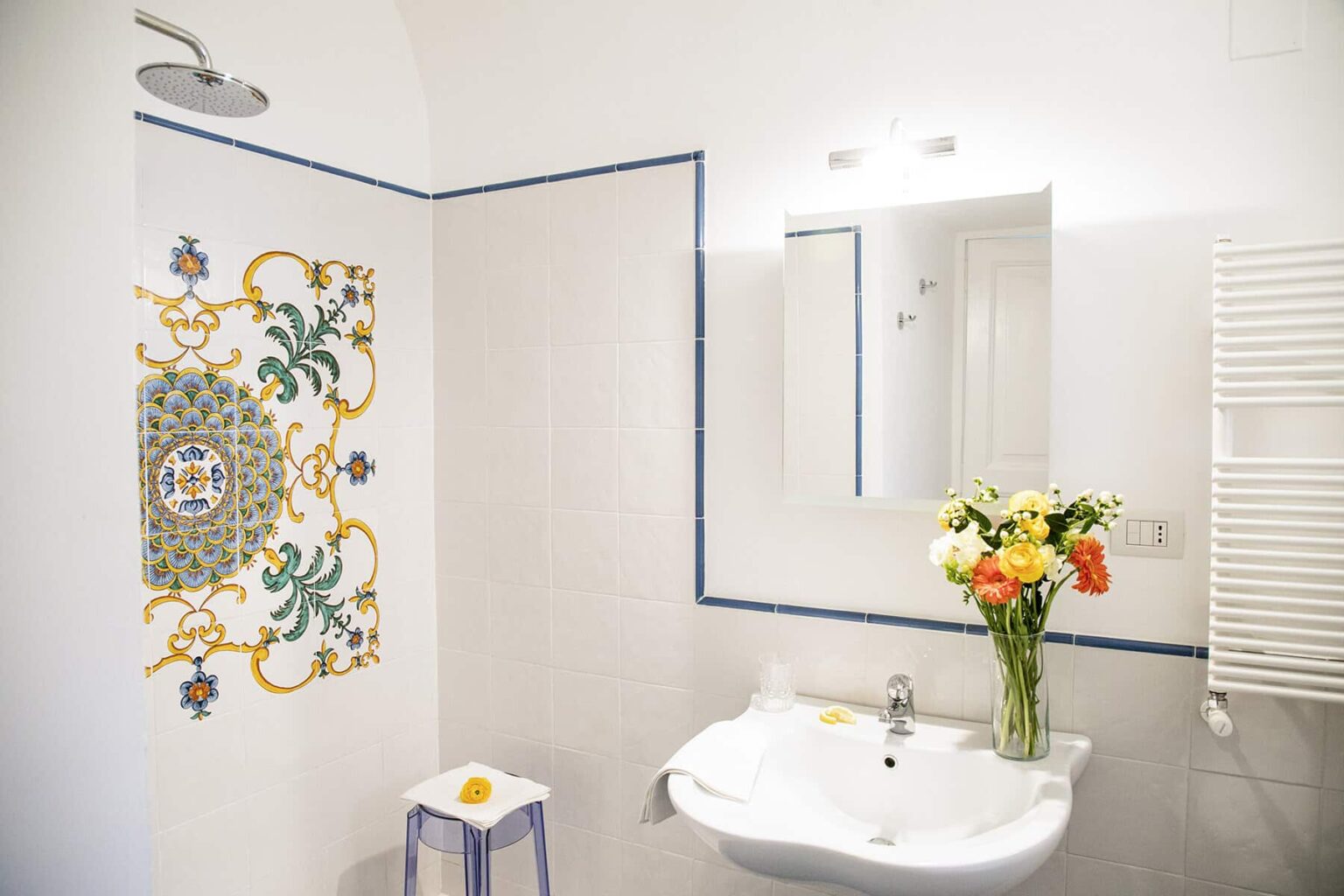 Bright and modern bathroom featuring elegant floral tiles on the wall, a sleek sink, a large mirror, and a vase of colorful flowers on the countertop. The space is well-lit and showcases a minimalist design with a touch of decorative charm. | Only on SopranoVillas