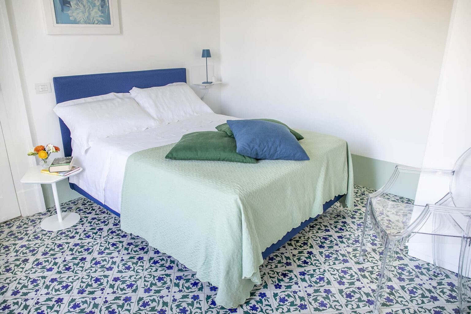 Cozy bedroom featuring a blue bed frame with white linens and green throw pillows, complemented by a floral patterned tile floor and a small bedside table with a vase of flowers. | Only on SopranoVillas