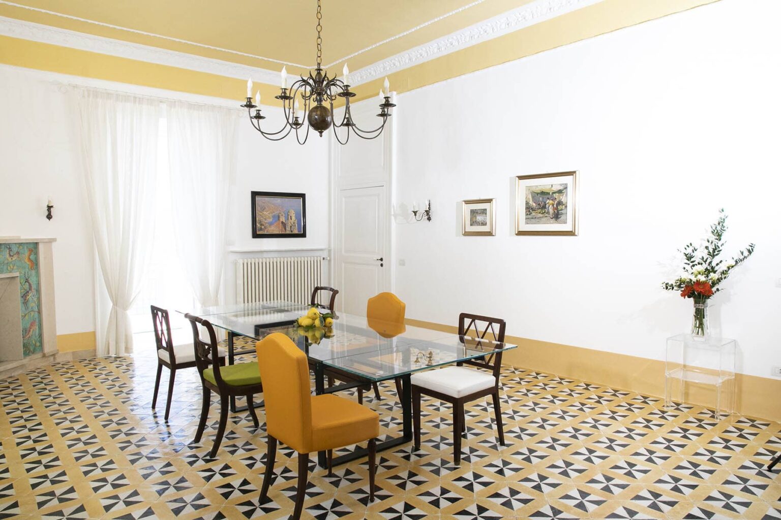 Bright and airy dining room featuring a glass dining table surrounded by six chairs, two of which are upholstered in orange. The room has a patterned tiled floor, elegant chandelier, and decorative artwork on the walls. A vase with flowers adds a touch of color to the space. | Only on SopranoVillas