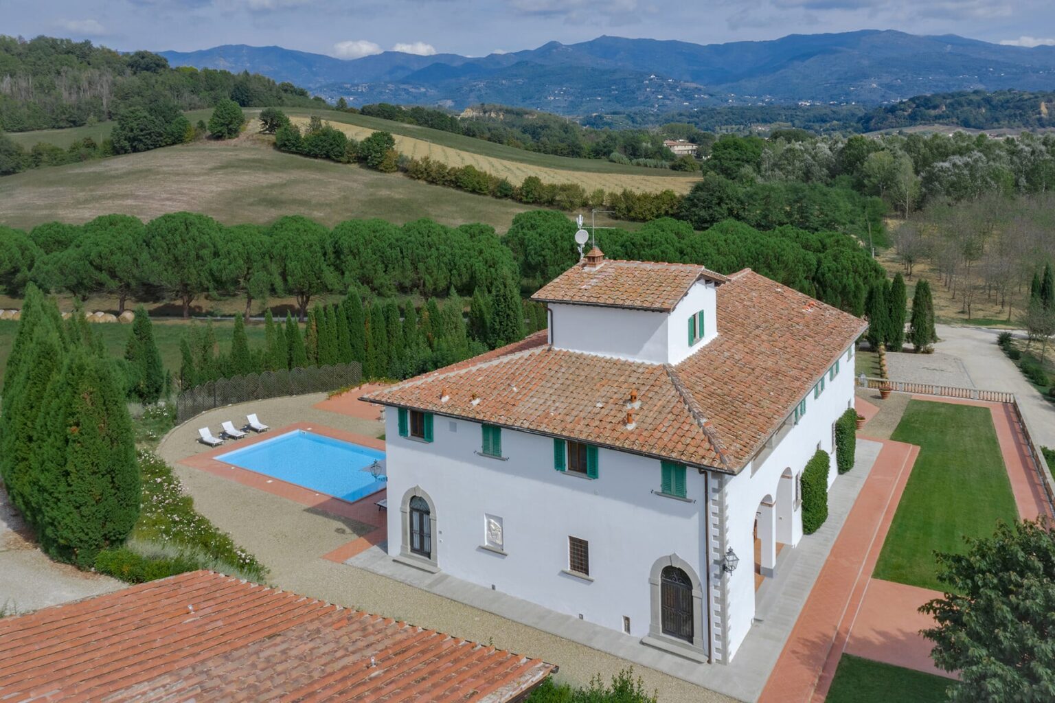 Aerial view of a spacious white villa with a terracotta roof, surrounded by lush greenery and rolling hills, featuring a blue swimming pool and well-maintained gardens. Ideal for luxury retreats or vacation rentals in a scenic countryside setting. | Only on SopranoVillas