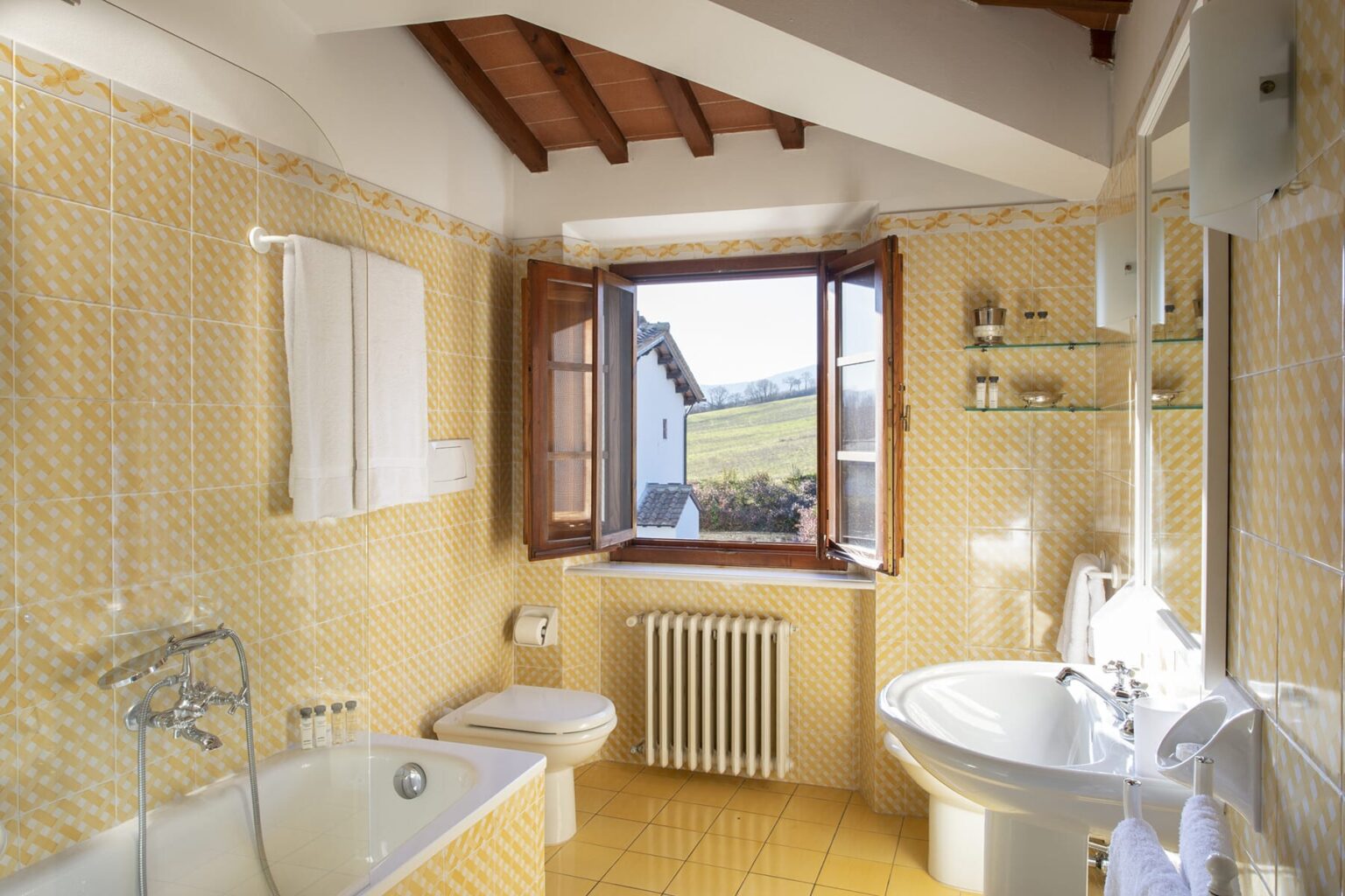 Bright and airy bathroom featuring a bathtub, modern sink, and large window with scenic views. The warm yellow tiles and wooden beams create a cozy atmosphere. Ideal for relaxation and comfort. | Only on SopranoVillas