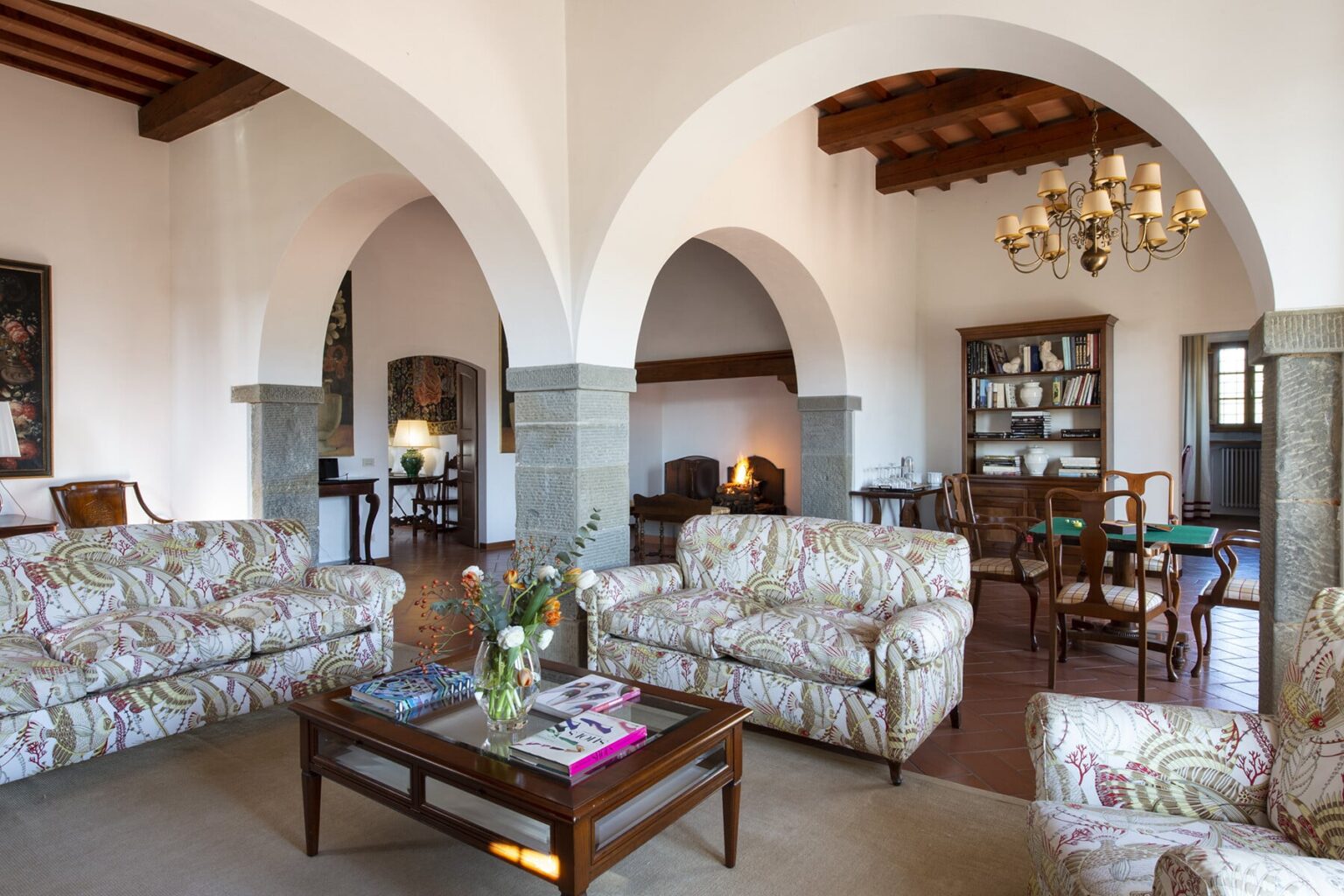Spacious living room with elegant floral sofas, wooden beams, and arched doorways, featuring a cozy dining area and a vintage bookshelf. | Only on SopranoVillas