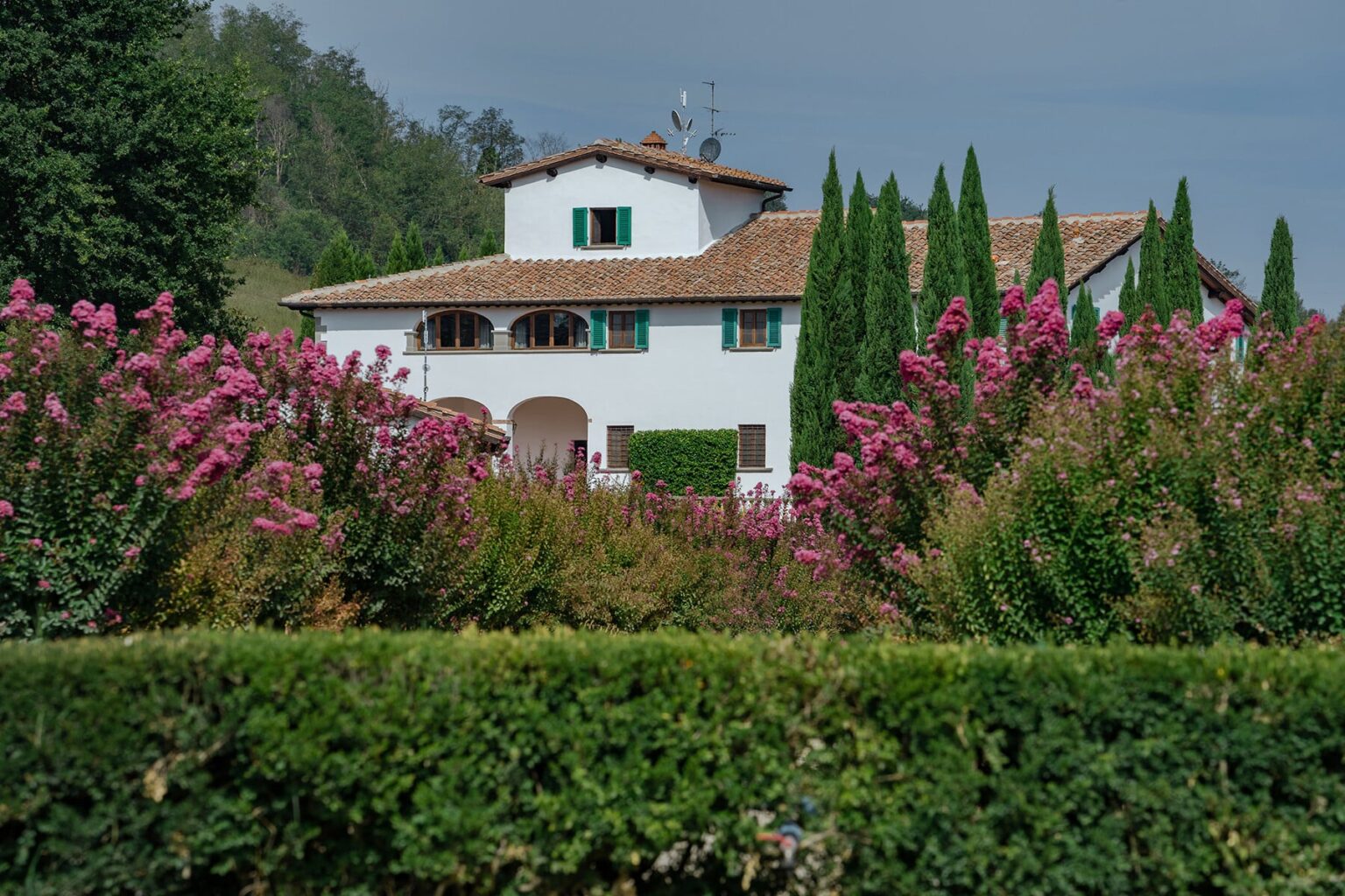 Charming white villa surrounded by vibrant pink flowers and lush greenery under a cloudy sky. Ideal for countryside retreats and nature photography. | Only on SopranoVillas