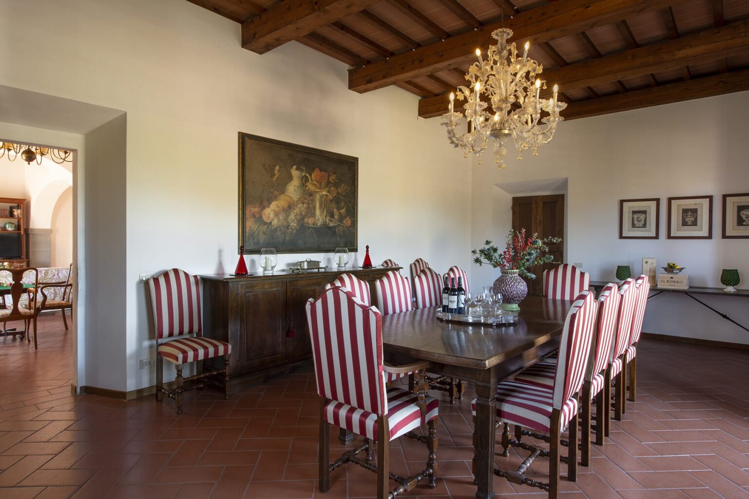 Elegant dining room featuring a large wooden table surrounded by striped chairs, a decorative chandelier, and artwork on the walls, creating a sophisticated atmosphere. | Only on SopranoVillas