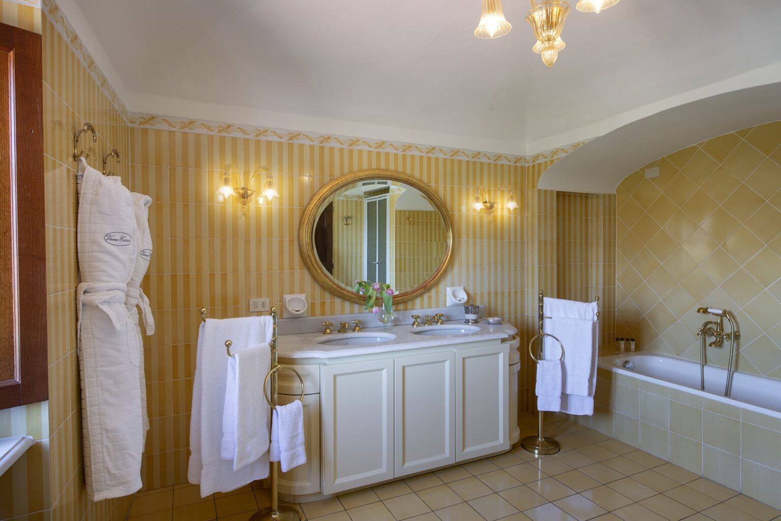 Luxurious bathroom featuring a large mirror, elegant lighting, a double sink vanity, and a freestanding bathtub. Soft neutral tones and plush towels create a serene atmosphere. | Only on SopranoVillas