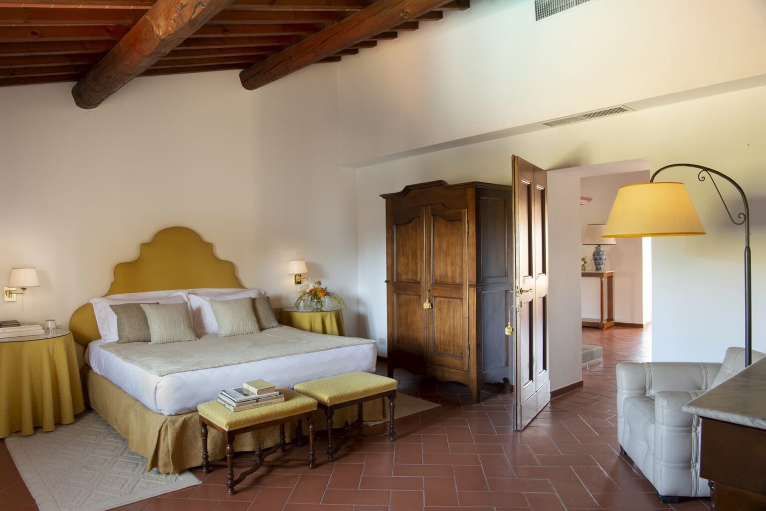 Spacious hotel room with a cozy bed, warm yellow accents, wooden beams, and stylish furniture, featuring a wardrobe, bedside lamps, and a comfortable seating area. Ideal for a relaxing stay. | Only on SopranoVillas