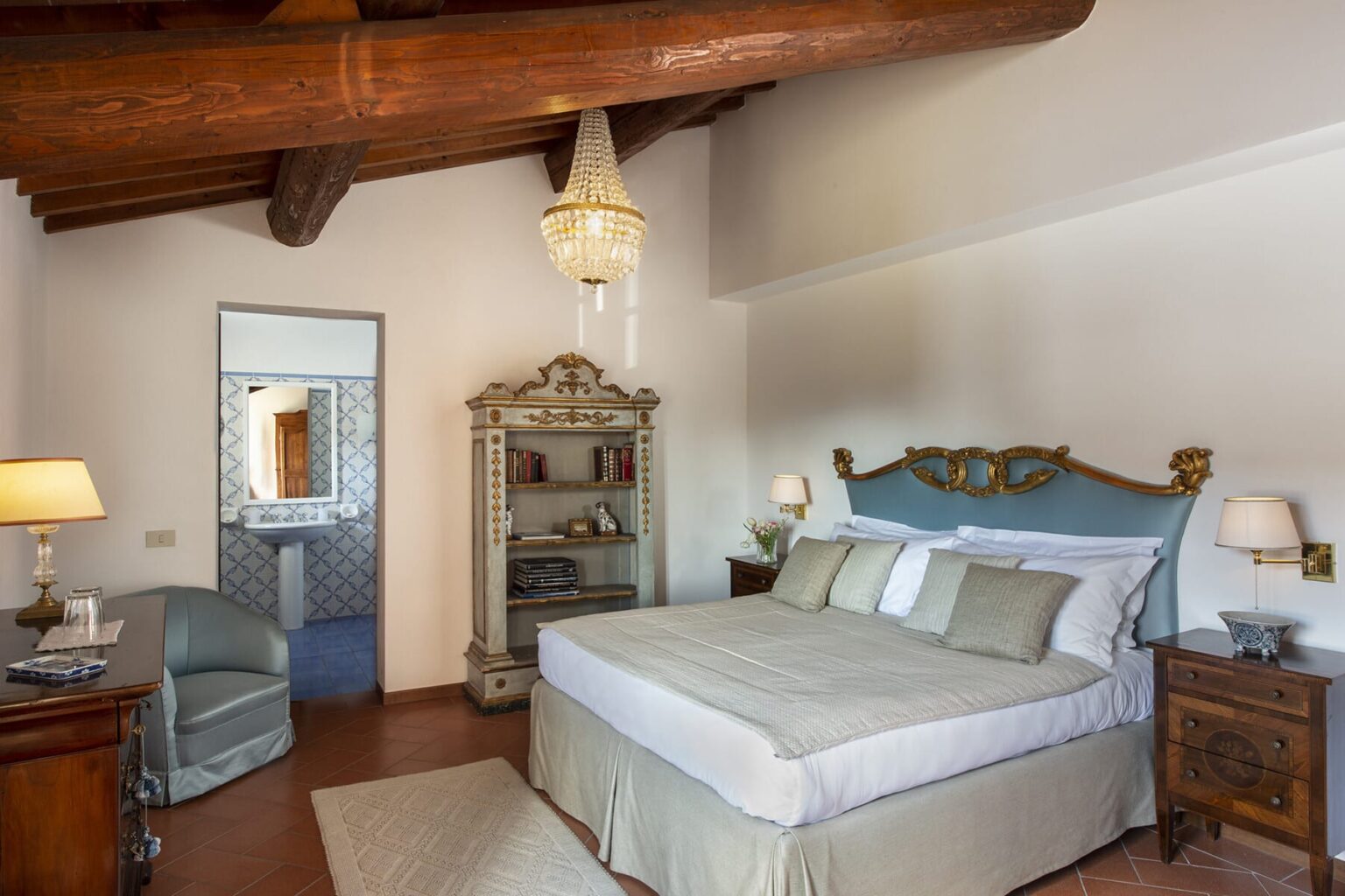 Elegant bedroom featuring a plush king-sized bed with decorative pillows, wooden beams on the ceiling, a vintage wardrobe, and soft lighting from a chandelier. The room has a cozy armchair and a mirror reflecting a stylish bathroom, creating a serene and inviting atmosphere. | Only on SopranoVillas