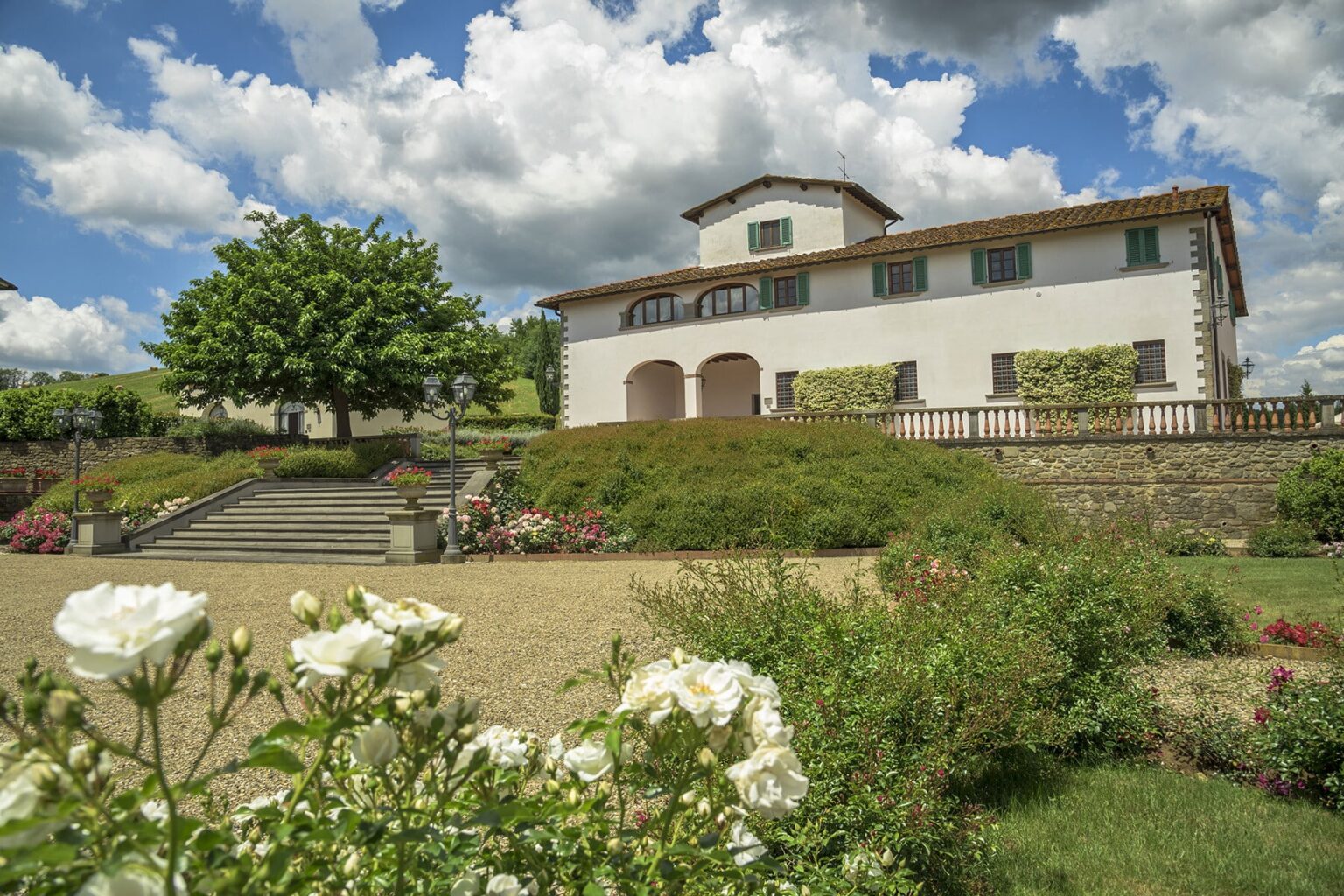 Charming countryside villa surrounded by lush gardens and blooming white roses, under a blue sky with fluffy clouds. Ideal for vacation rentals or destination weddings. | Only on SopranoVillas