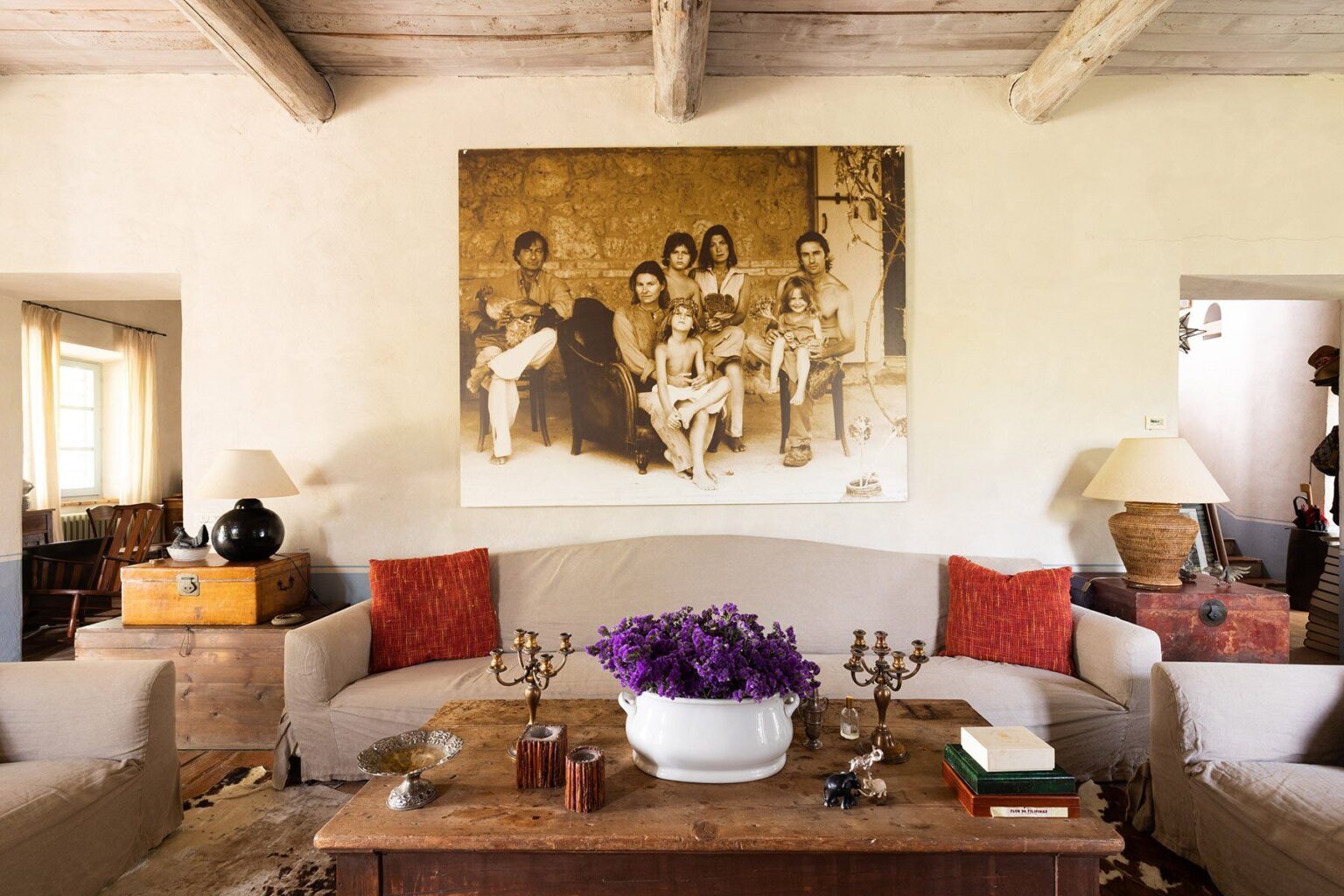 Cozy living room featuring a large art piece of a family portrait on the wall, a plush beige sofa with red cushions, a wooden coffee table adorned with a white vase of purple flowers, and warm lighting from table lamps. | Only on SopranoVillas