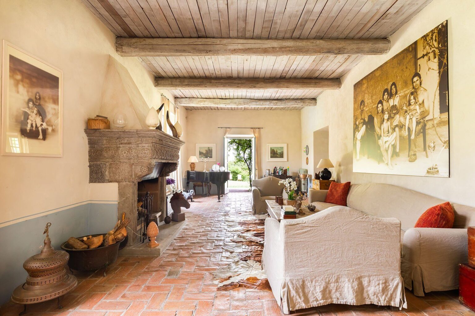 Interior view of a cozy living room featuring rustic wooden beams, a stone fireplace, and natural light flowing in from a doorway. The room is decorated with a mix of modern and vintage art pieces, comfortable furniture, and warm color tones, creating an inviting atmosphere. | Only on SopranoVillas