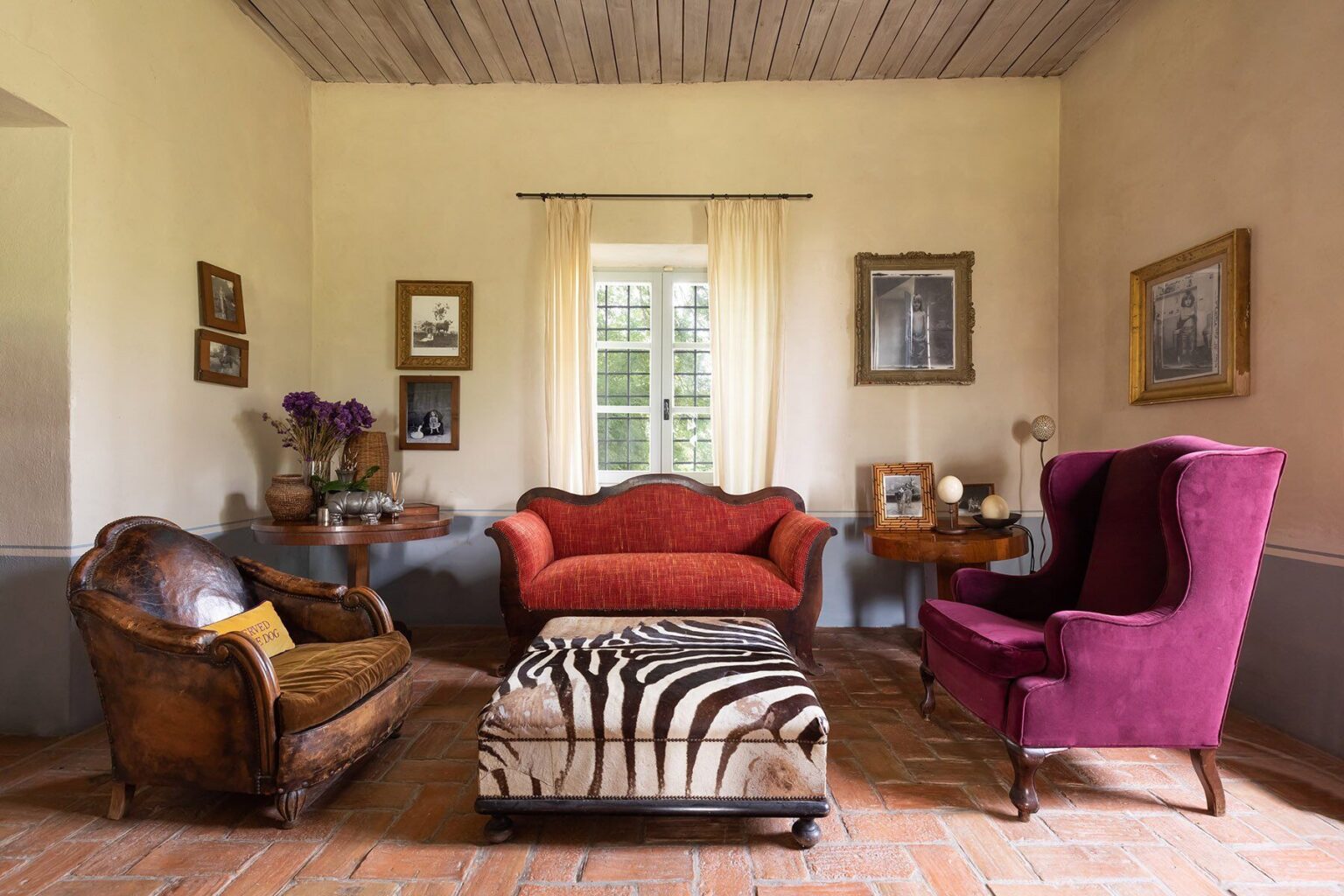 Cozy living room interior featuring a vintage brown leather sofa, a vibrant red loveseat, and a purple wingback chair. A zebra-striped ottoman sits in the center, surrounded by framed artwork on the walls and a window with sheer curtains allowing natural light to illuminate the space. | Only on SopranoVillas