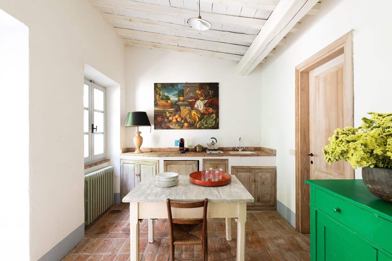 Bright and airy kitchen featuring a wooden table with chairs, a green cabinet, and a wall-mounted painting of flowers. Natural light streams in through the window, enhancing the warm, inviting atmosphere. | Only on SopranoVillas